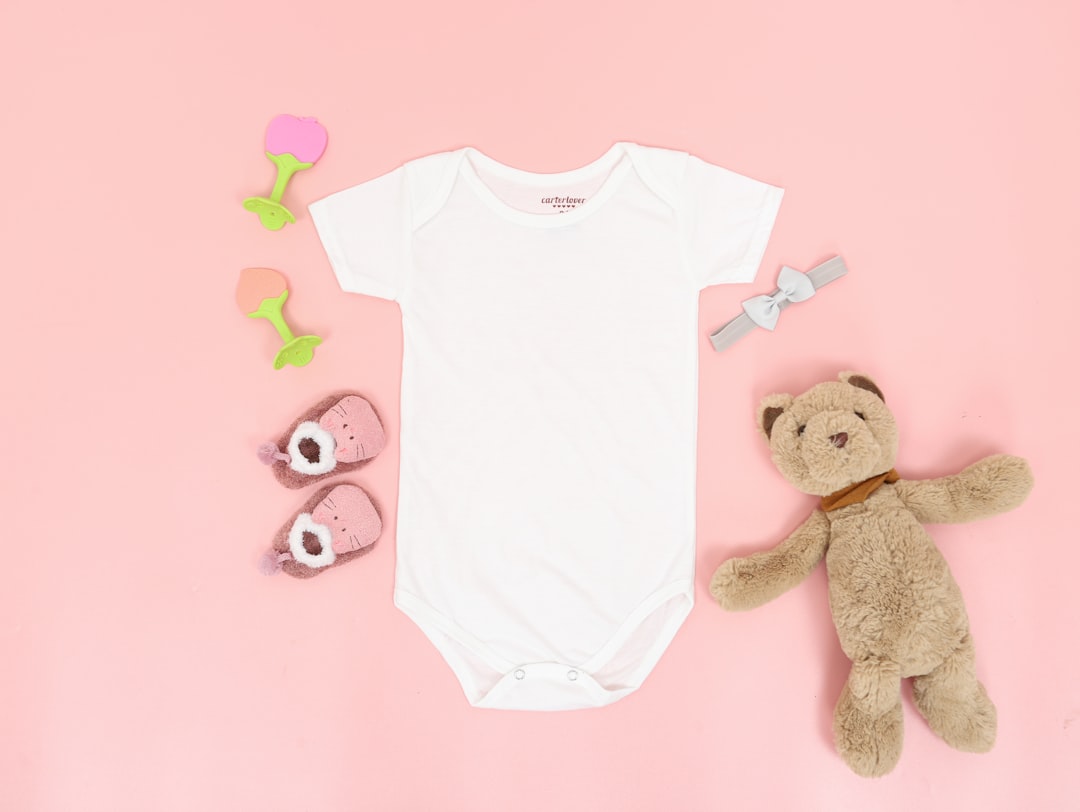 Home A onesie lying on a pink surface next to baby toys and a cute teddy bear.